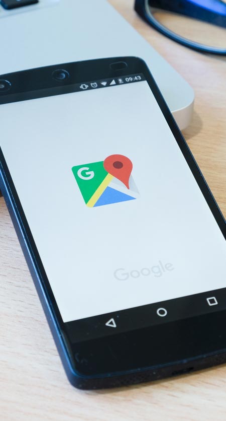 Auf einem hellen Holztisch neben einem Computer-Trackpad und einem schwarzen Kabel liegt ein Smartphone, das den Ladebildschirm der Google Maps-App anzeigt und Tools für lokale Suchmaschinenoptimierung hervorhebt.